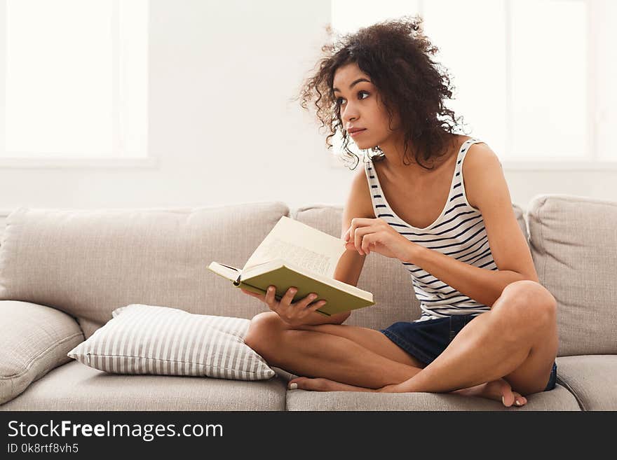 Thoughtful african-american student girl reading book. Young woman studying at home, sitting on beige couch, copy space. Thoughtful african-american student girl reading book. Young woman studying at home, sitting on beige couch, copy space