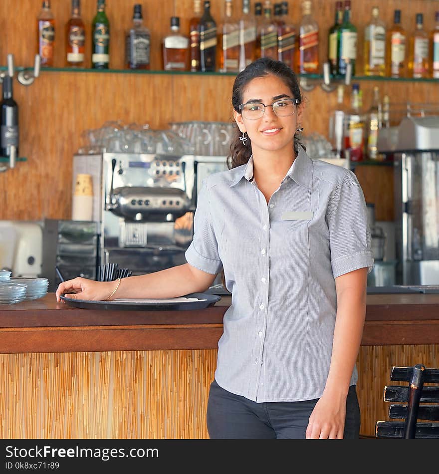 The waitress is standing near the restaurant bar of the hotel. A waitress with a tray is waiting for the order. Shelves with bottles of alcohol in the background. The concept of service. The waitress is standing near the restaurant bar of the hotel. A waitress with a tray is waiting for the order. Shelves with bottles of alcohol in the background. The concept of service.