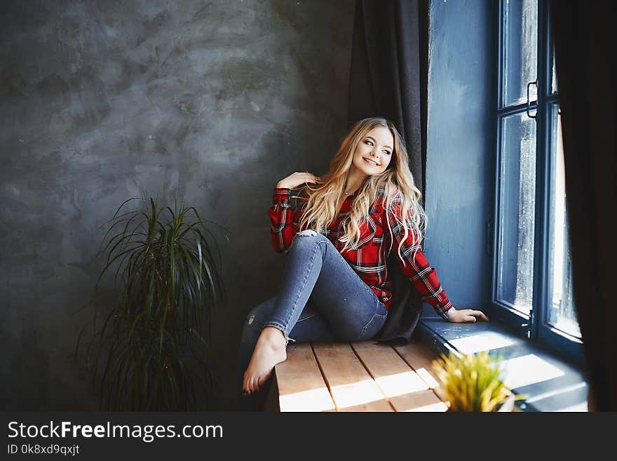 Happy And Smiling Beautiful Blonde Girl In Jeans And Red Plaid Shirt, Sits And Posing Near The Window And Looks Outside