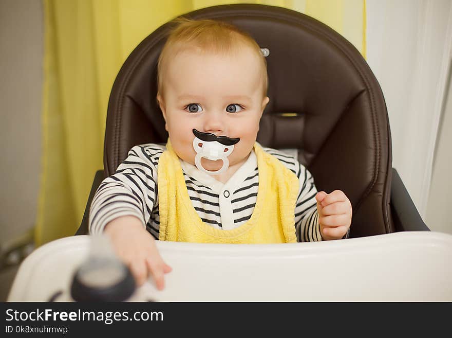 Little funny boy sitting in a chair for feeding with childish nipple in the form of a mustache. Indoor