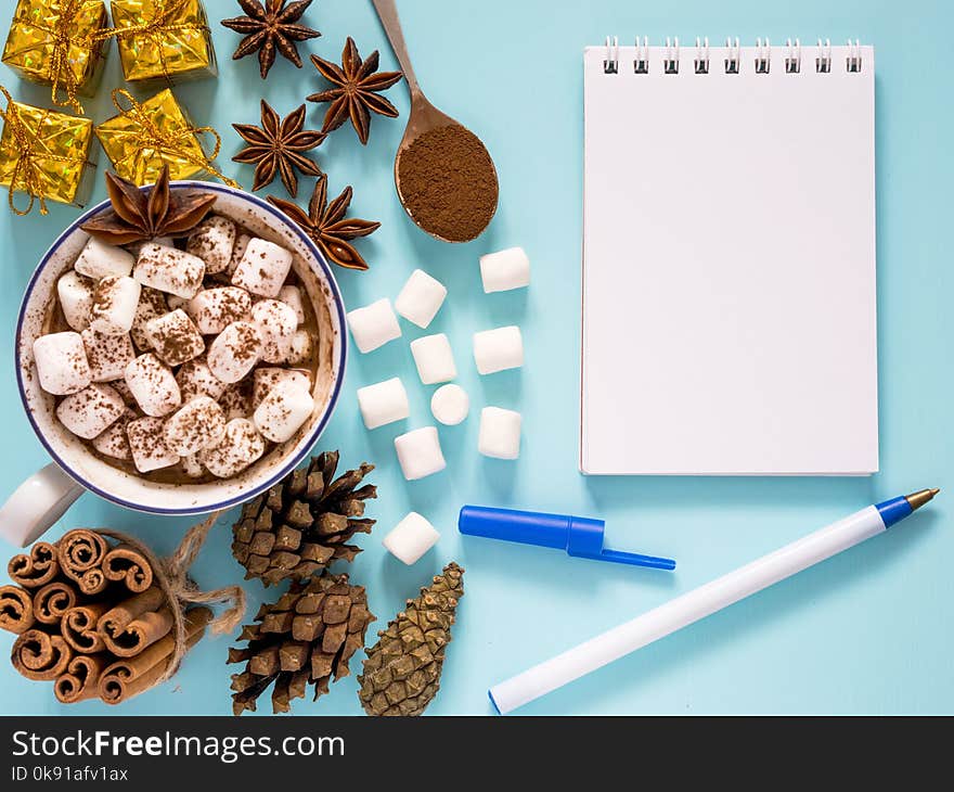 Cup of hot cocoa or carob or chocolate with marshmallow and winter spices star anise and cinnamon sticks, pine cones and carob powder in spoon and gifts and empty notebook on turquoise table. Flat lay. Cup of hot cocoa or carob or chocolate with marshmallow and winter spices star anise and cinnamon sticks, pine cones and carob powder in spoon and gifts and empty notebook on turquoise table. Flat lay