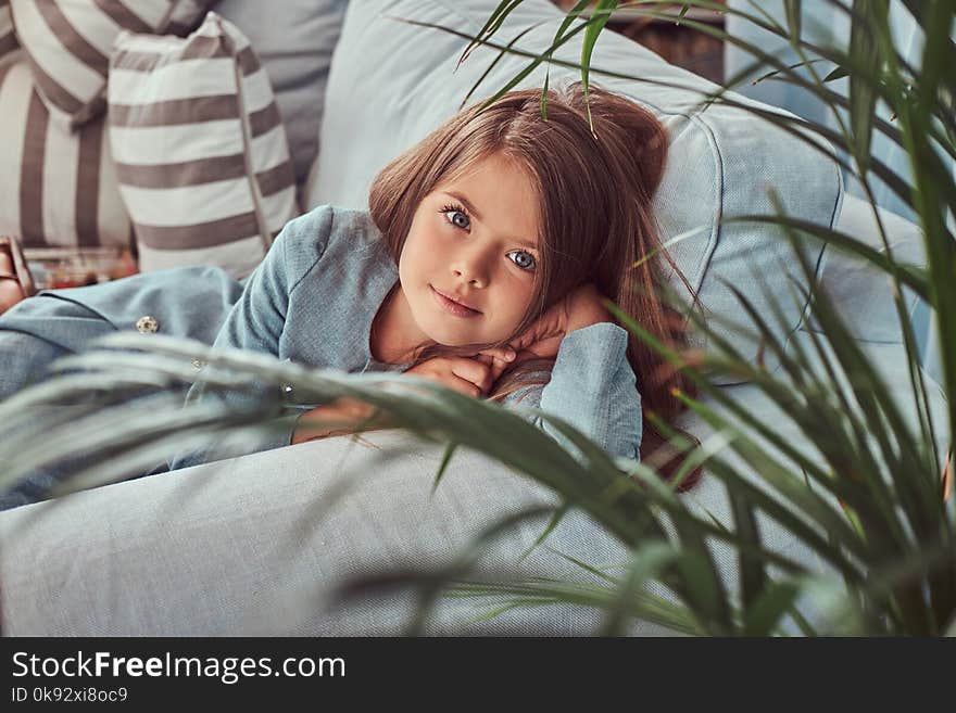Portrait of a cute little girl with long brown hair and piercing glance, looking at a camera, lying on a sofa at home alone.