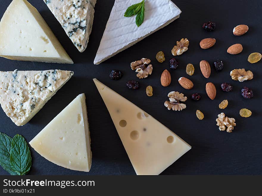 Different kinds of cheeses on black stone board with nuts and herbs.