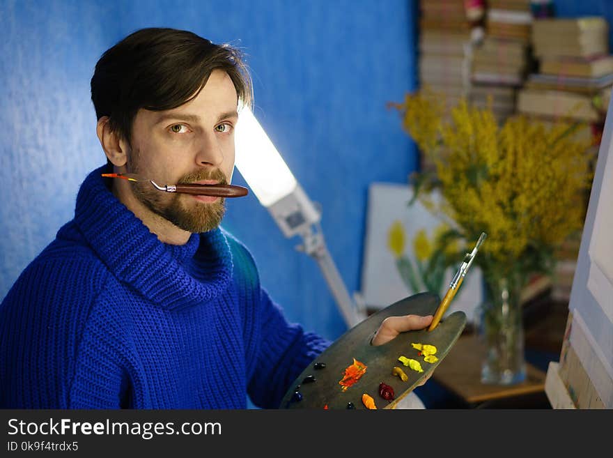 Artist holds a palette in his mouth. He holds palette with oil paints and brushes. Canvas and flowers in background. Artist holds a palette in his mouth. He holds palette with oil paints and brushes. Canvas and flowers in background