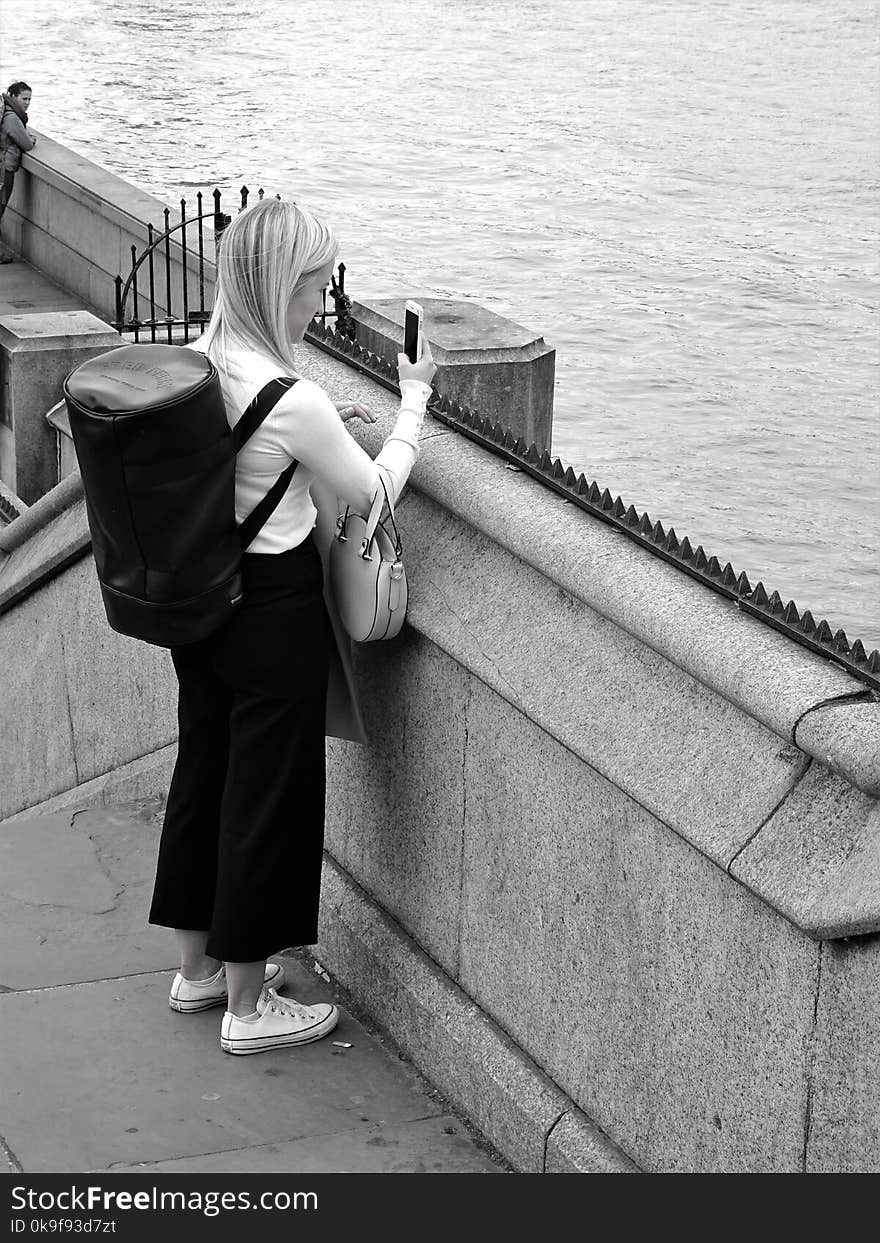 Black and White Photo of Woman Holding Smartphone While Standing Near Calm Water