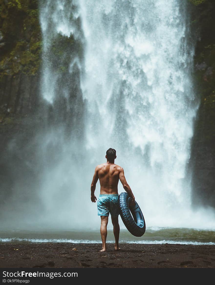 Man Wearing Blue Shorts Holding Vehicle Tire Facing Waterfalls