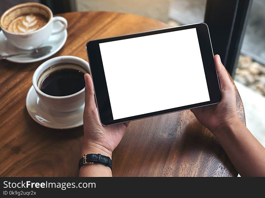 Mockup image of hands holding black tablet pc with white blank screen and coffee cups on table