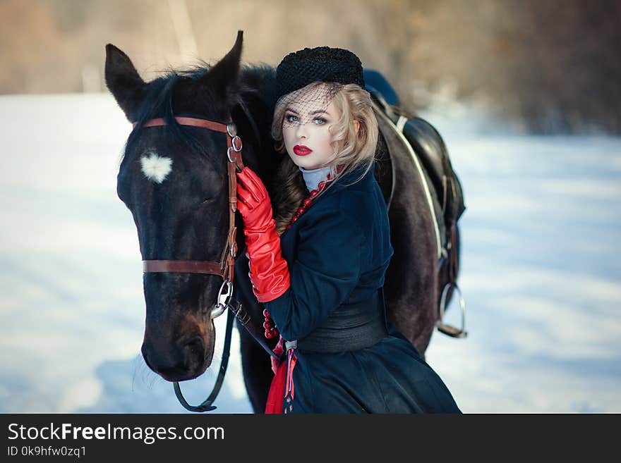 Blond woman in red dress with long black coat with dark horse on the background of winter landscape. Blond woman in red dress with long black coat with dark horse on the background of winter landscape