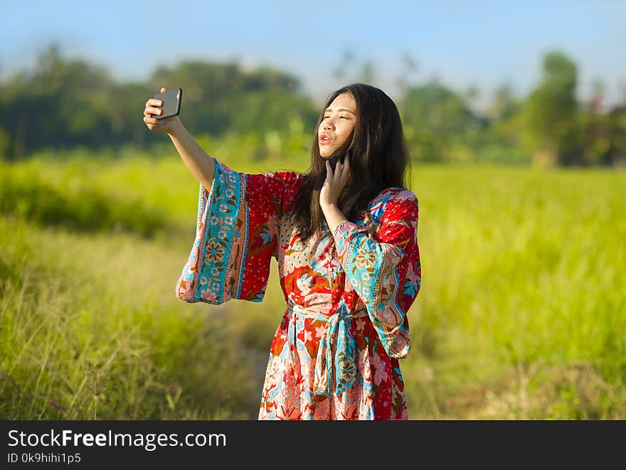 Young beautiful and happy Asian Chinese tourist woman on her 20s with colorful dress taking selfie pic with mobile phone camera on tropical green grass field background in holidays and travel concept