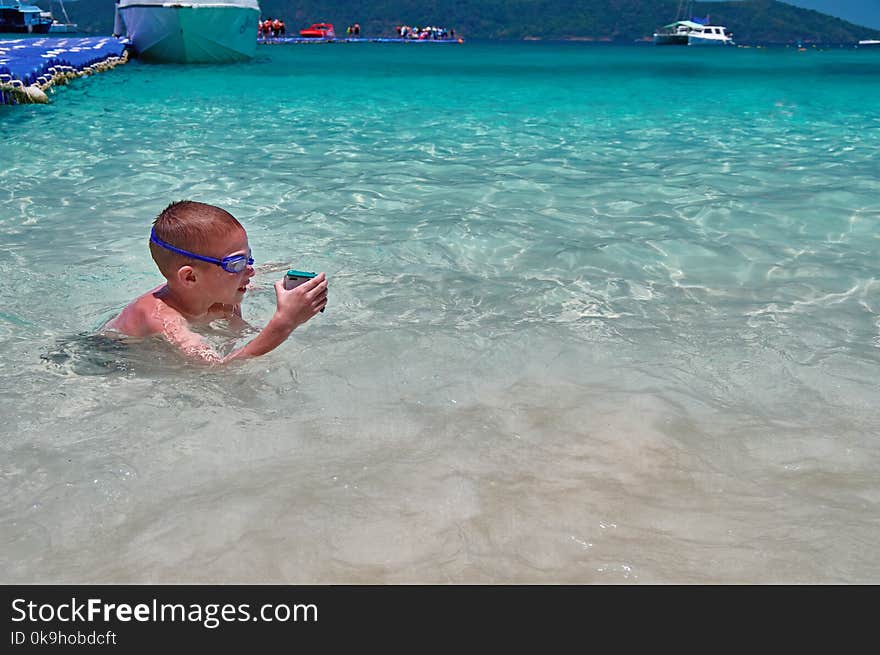 Little boy in swimming glasses plays in turquoise water with action camera in protective box. Child swims in waters tropical sea.