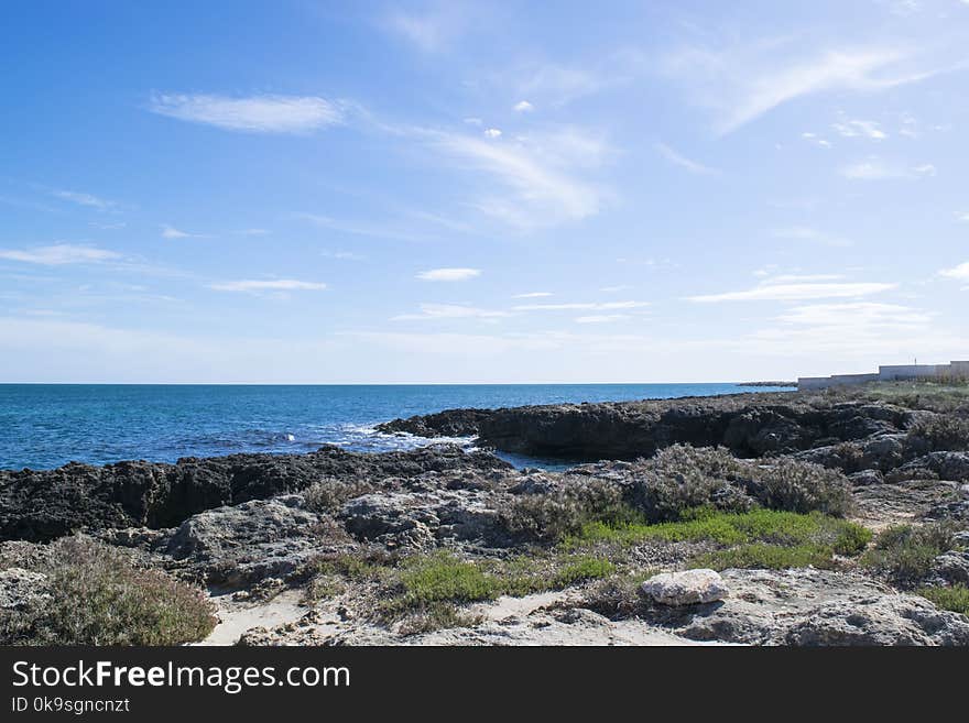 Wavy Sea Beside Rock Formations Near Sea Shore With Grasses Under Blue Cloudy Sky at Daytime