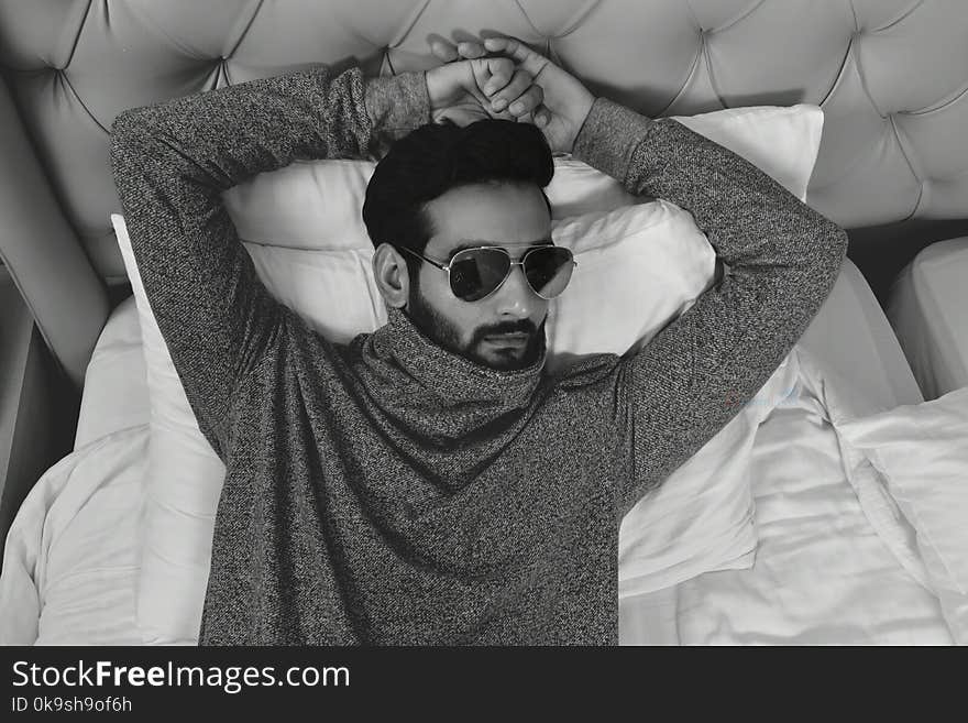 Grayscale Photo of Man in Long-sleeved Wearing Sunglasses Lying on Bed With Both Hand on Head