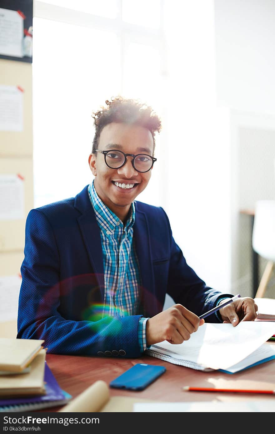 Happy student looking at camera with smile while sitting by workplace with paper