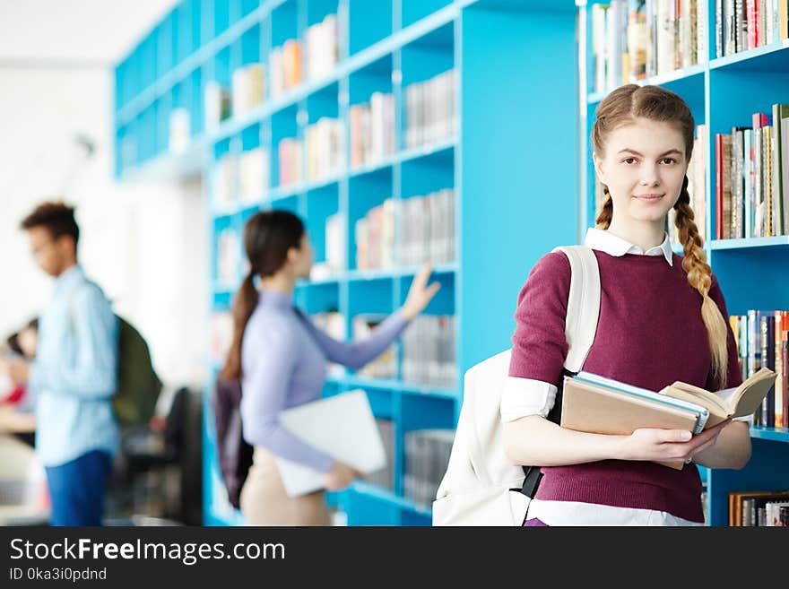 One of students with several books looking at camera in library on background of her groupmates. One of students with several books looking at camera in library on background of her groupmates