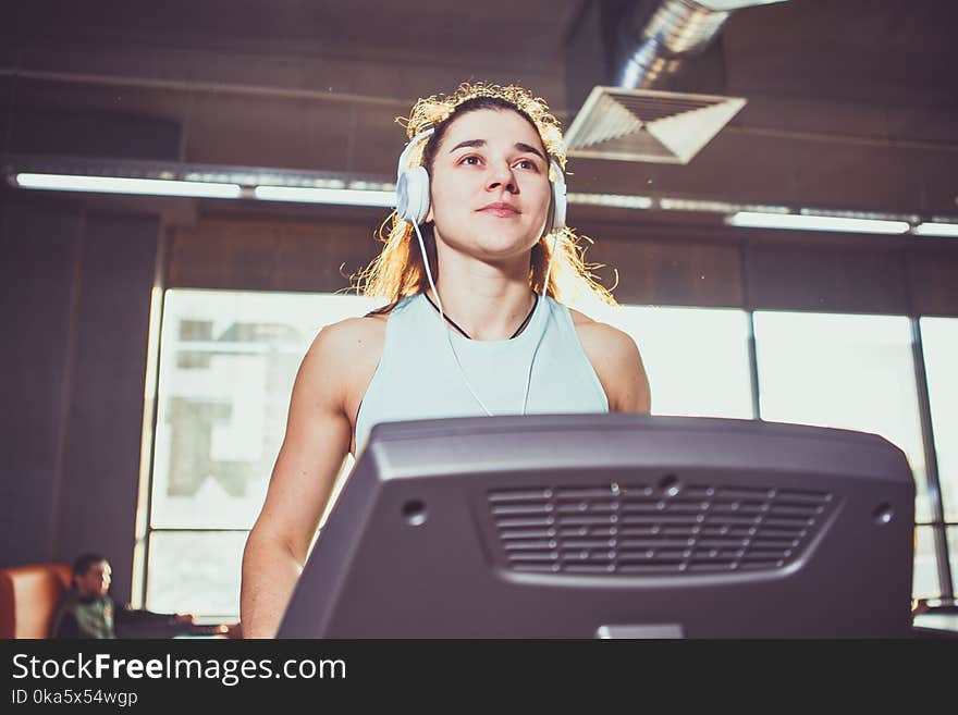 Theme is sport and music. A beautiful inflated Caucasian woman running in a gym on a treadmill. On her head are big white headphones, the girl listens to music during a cardio workout for weight loss. Theme is sport and music. A beautiful inflated Caucasian woman running in a gym on a treadmill. On her head are big white headphones, the girl listens to music during a cardio workout for weight loss.