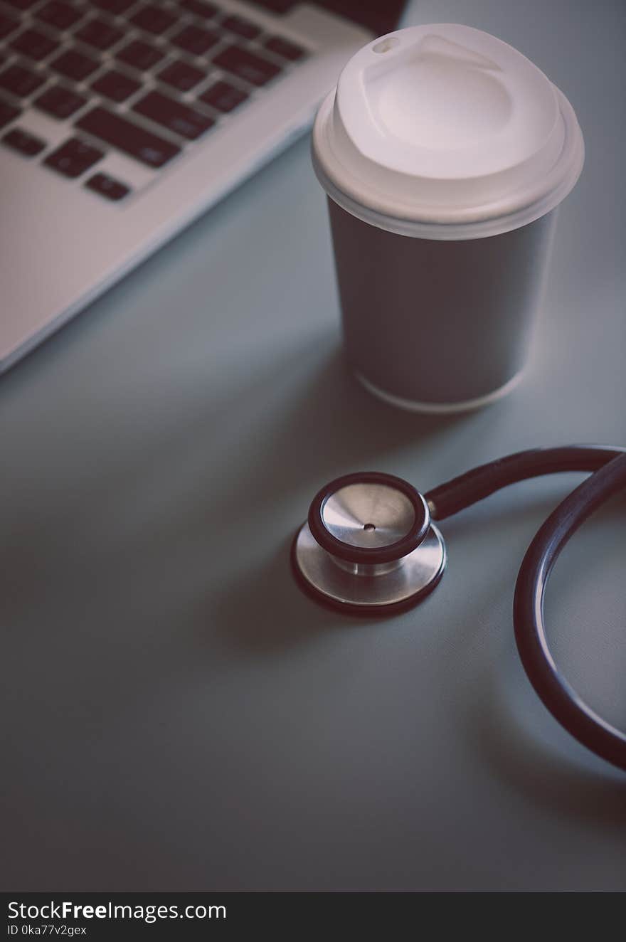 Gray and Black Stethoscope Beside Disposable Cup and Macbook Pro