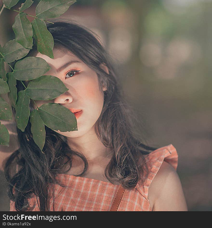 Selective Focus Photography of Woman in Orange Sleeveless Top Hiding Face Behind Tree&#x27;s Leaf