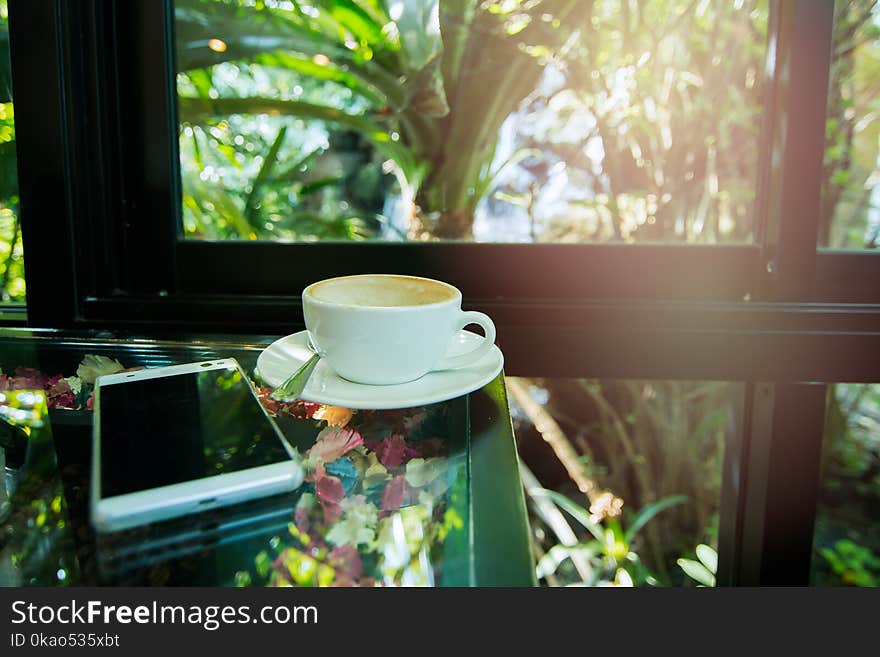 Picture of a white cup of coffee by the window. There is a smartphone next to it.At cafe