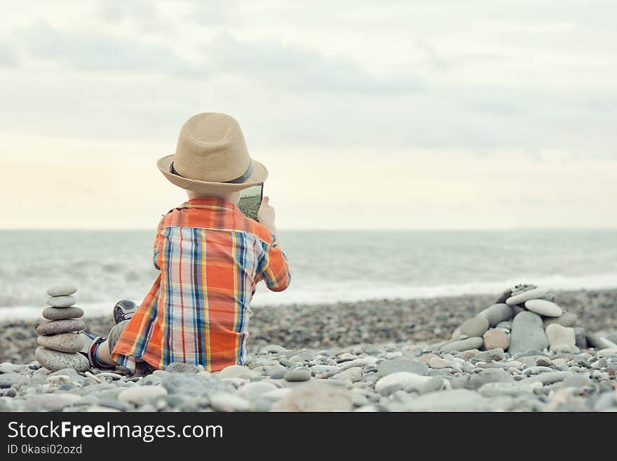 Little boy takes pictures on the smart phone. Sits on a pebble beach.