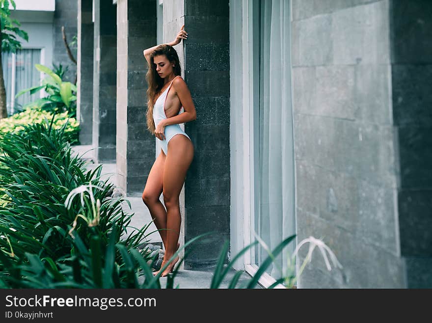 Beautiful brunette girl in white swimsuit posing in tropical location with green trees. Young sports model in bikini