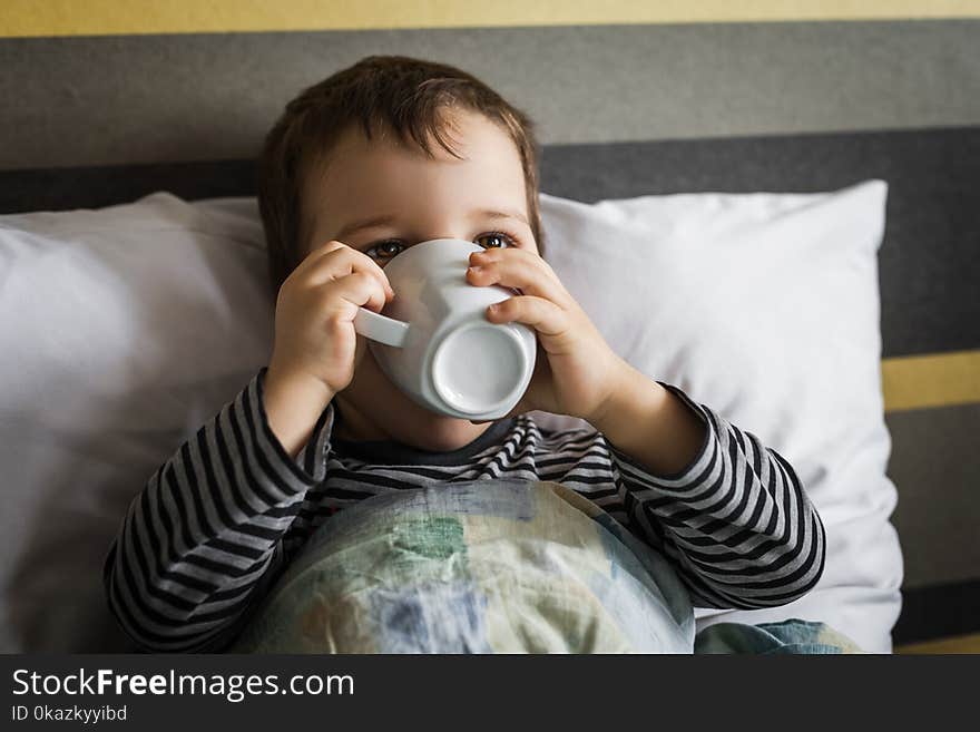 Unhealthy little boy lying on bed on pillow under blanket drinking medicine syrup in cup. Unhealthy little boy lying on bed on pillow under blanket drinking medicine syrup in cup