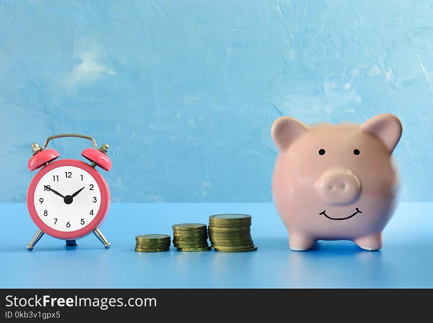 On a dark blue background photographed piggy bank, next to her a small pink alarm clock and three stacks of coins. Preservation of savings. A bright picture.