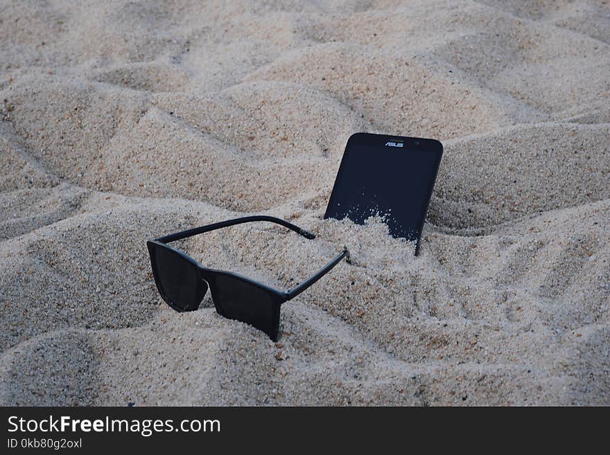 Black Wayfarer-style Sunglasses Beside Black Asus Android Smartphone on Brown Sand