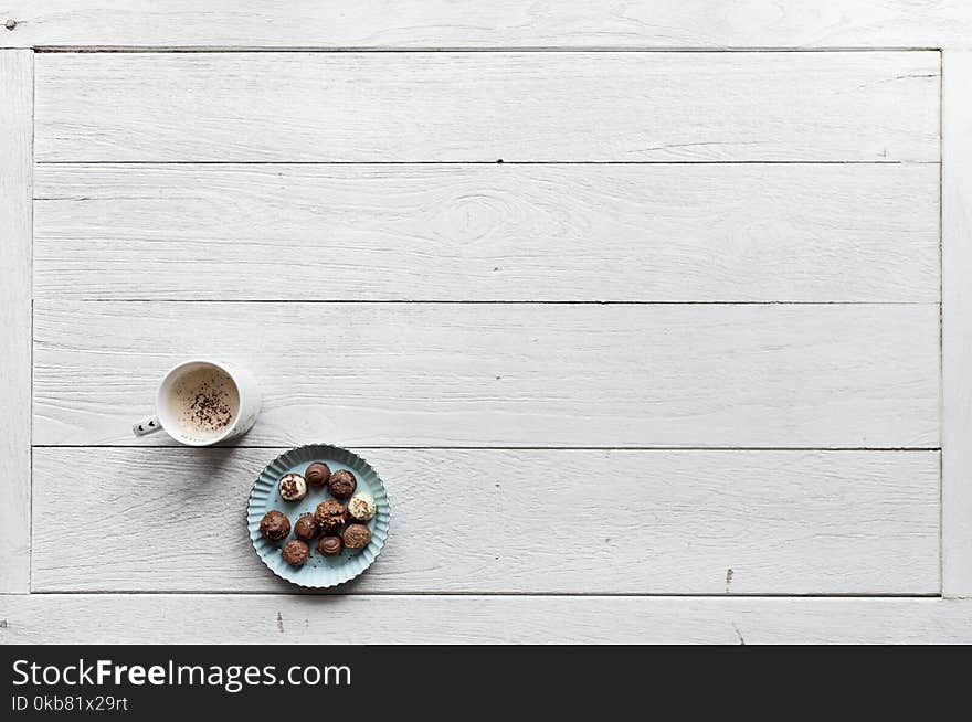 White Mug Filled With Coffee Beside Baked Pastries on Paper Plate All on Top of White Wood Surface