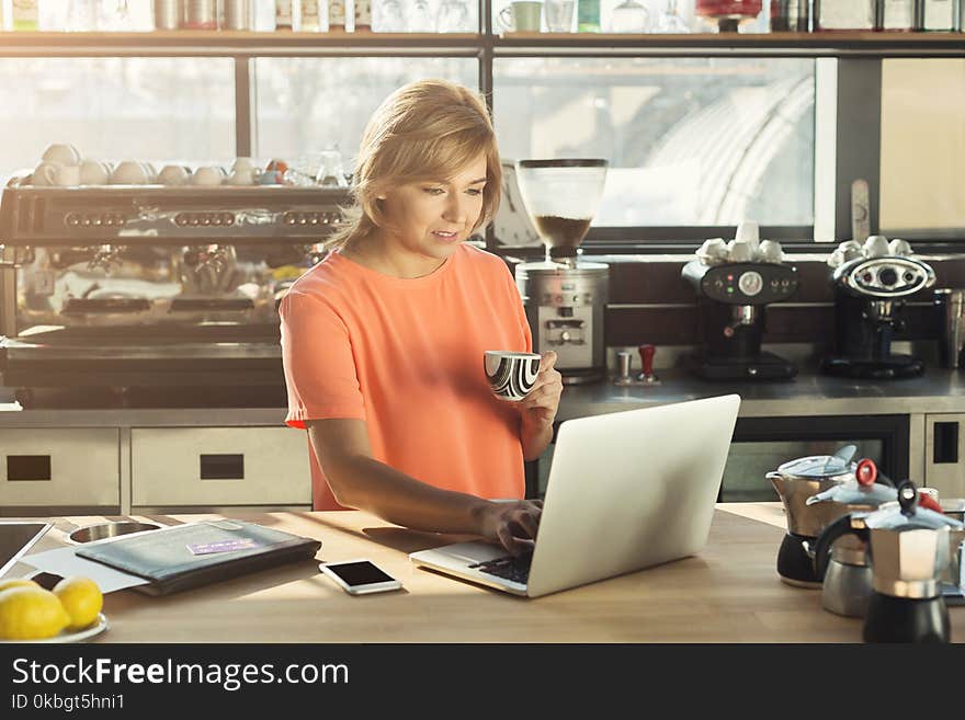 Middle-aged woman barista or manager working on laptop, making an order online and drinking coffee at modern cafeteria. Work, lifestyle, cafe, restaurant concept. Middle-aged woman barista or manager working on laptop, making an order online and drinking coffee at modern cafeteria. Work, lifestyle, cafe, restaurant concept