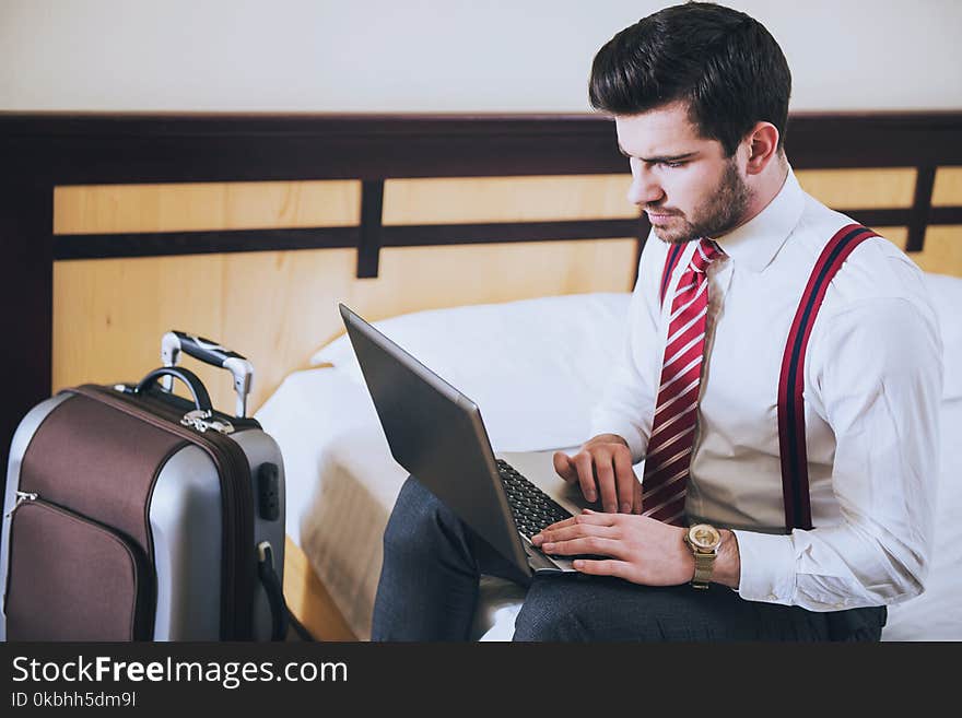 Man is sitting on bed in suit with suitcase in bedroom, working on laptop. Man is sitting on bed in suit with suitcase in bedroom, working on laptop.