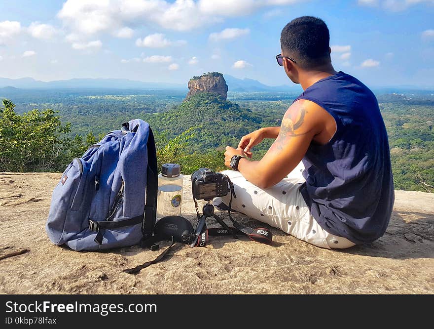 Man Sitting on Top of Gray Cliff Mountain Beside Backpack, Water Bottle, and Camera