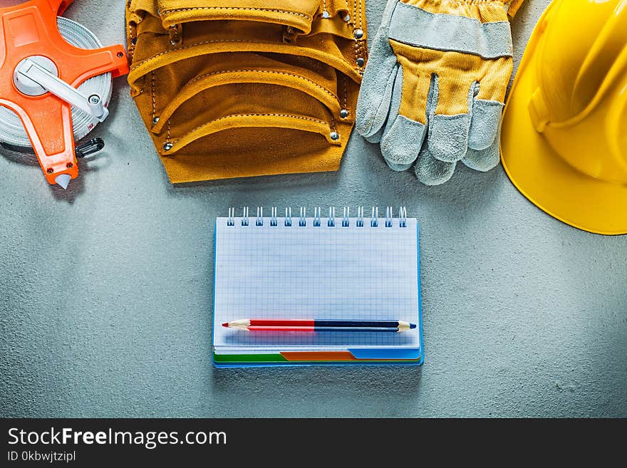 Protective gloves hard hat notepad pencil tool belt tape measure on concrete background.