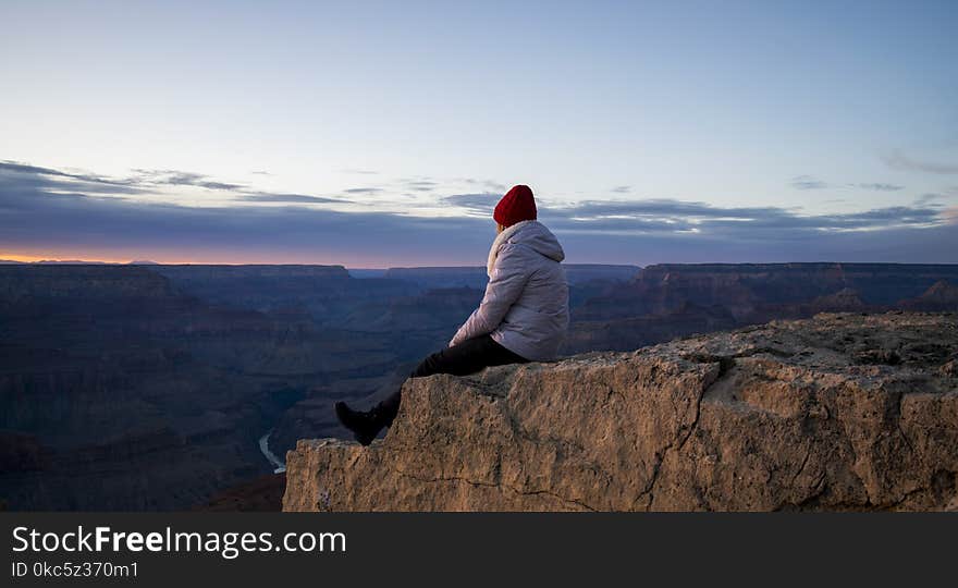 Person Wearing Gray Hooded Jacket and Black Pants Sitting on Mountain Cliff during Sunset