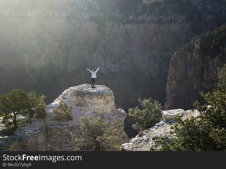 Woman in White Coat and Black Pants Standing on Brown Rock Formation Mountain