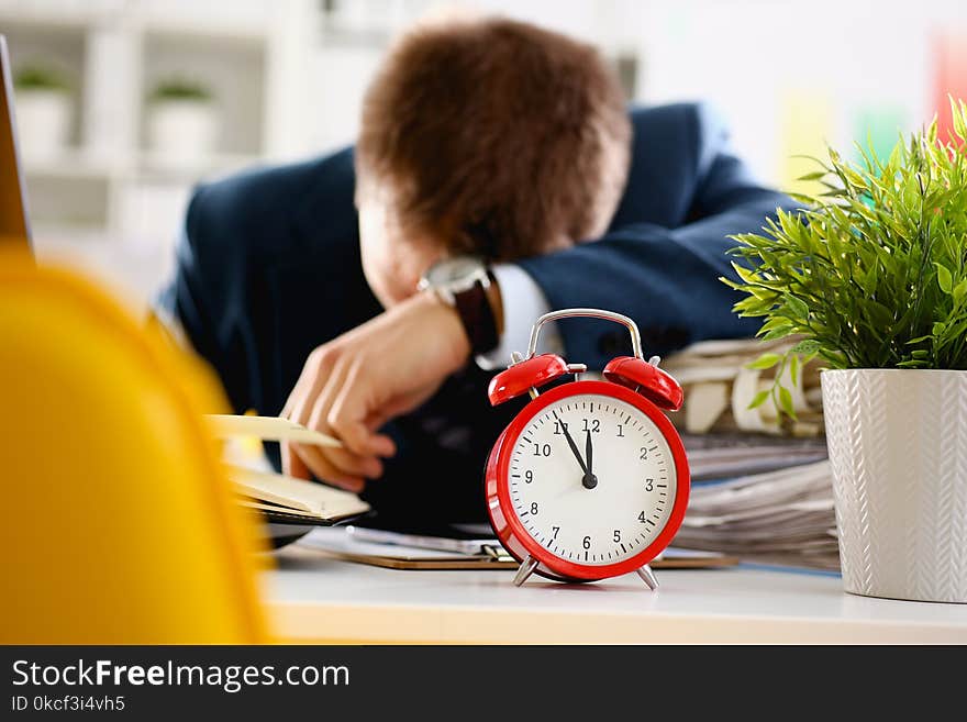 Red alarm clock shows late time closeup and tired office male clerk in suit take nap on table workplace full of exam papers. Career frustration freelance employment fail study problem low energy down