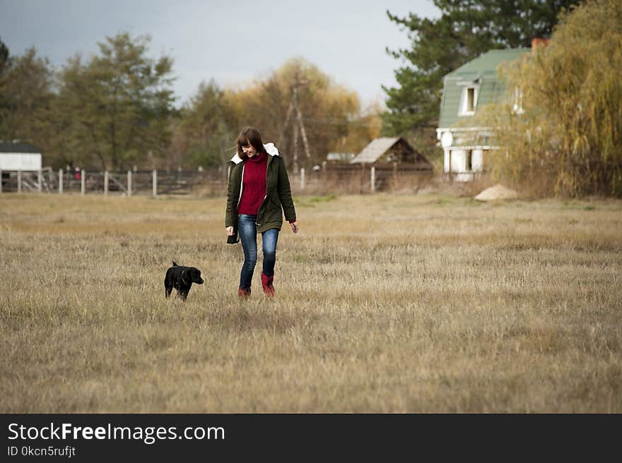 Woman Wearing Brown Zip-up Jacket and Blue Denim Jeans Walking Beside Short-coated Black Dog at Daytime