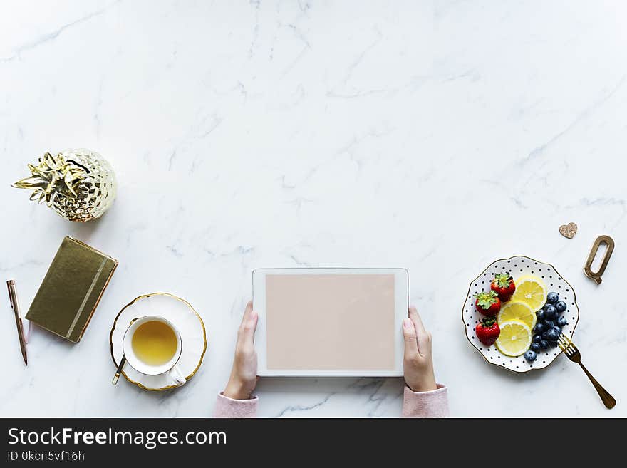 Person Holding White Tablet Computer Beside Teacup and Saucer of Blueberries