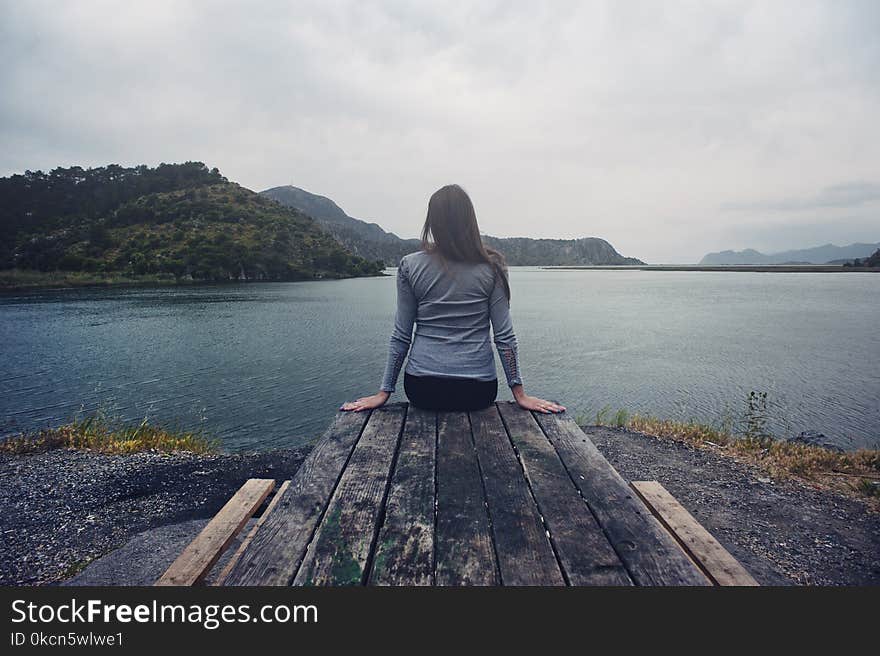 Woman Wearing Gray Long-sleeved Shirt and Black Black Bottoms Outfit Sitting on Gray Wooden Picnic Table Facing Towards Calm Body of Water at Daytime