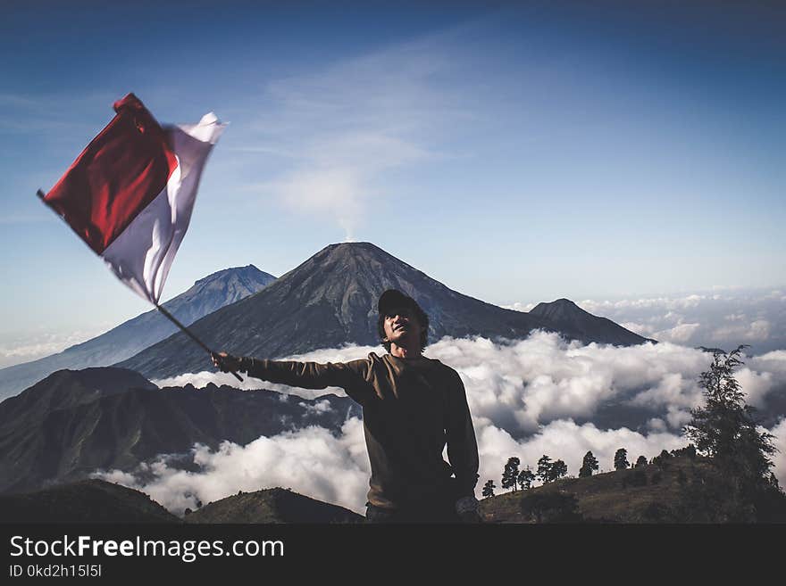 Man Wearing Black Crew-neck Sweater Holding White and Red Flag Standing Near Mountain Under Blue and White Sky