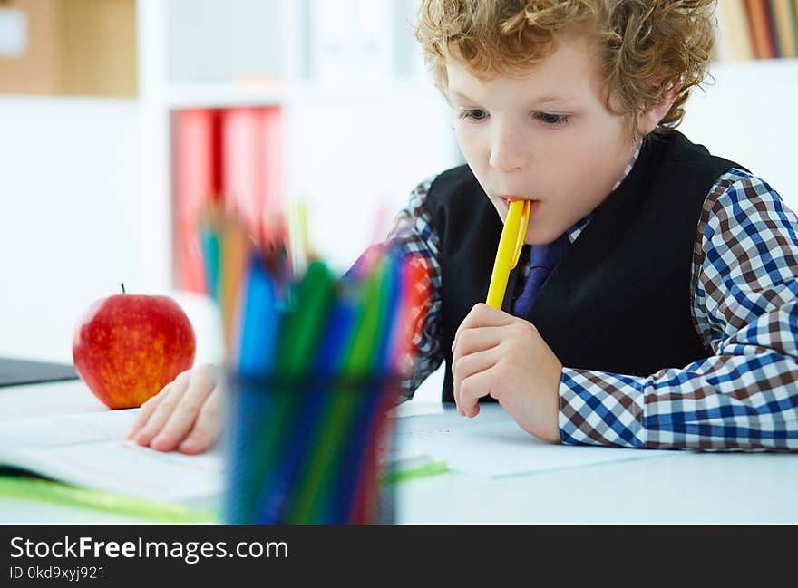 Curly Caucasian boy thought about the task and took a pen in his mouth during the lesson at school. Education, childhood