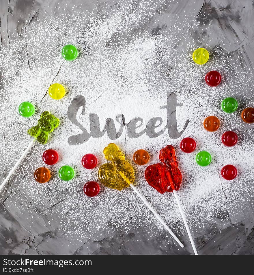 The word sweet is written on a gray concrete background with the help of powdered sugar next to multi-colored sweets and retro candies in the form of cockerels. Top view, flat lay. The word sweet is written on a gray concrete background with the help of powdered sugar next to multi-colored sweets and retro candies in the form of cockerels. Top view, flat lay