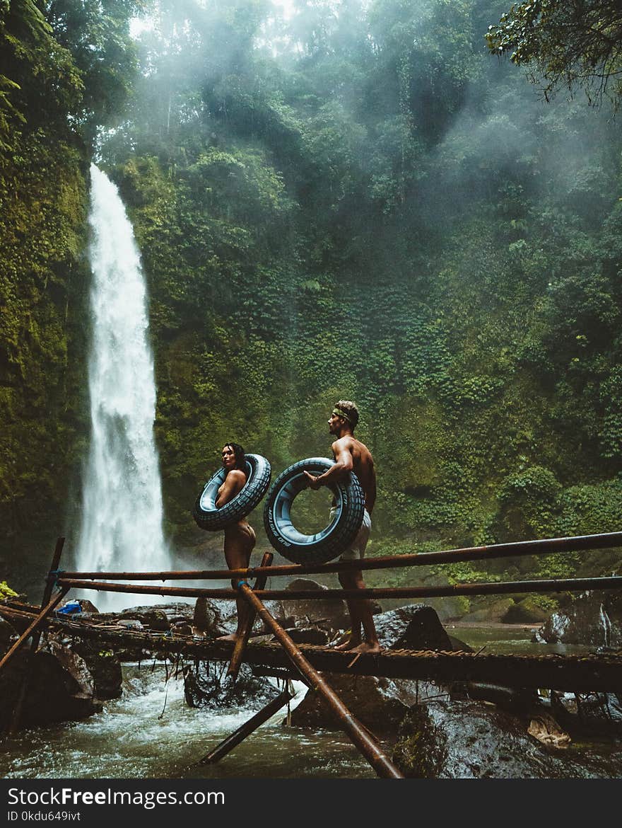 Two Person Carrying Black Inflatable Pool Float on Brown Wooden Bridge Near Waterfalls