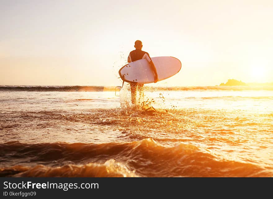 Surfer runs with surfboard towards ocean waves ta sunset time