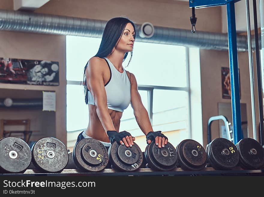 A beautiful athletic brunette female in a sportswear standing near the counter with dumbbells in the gym.