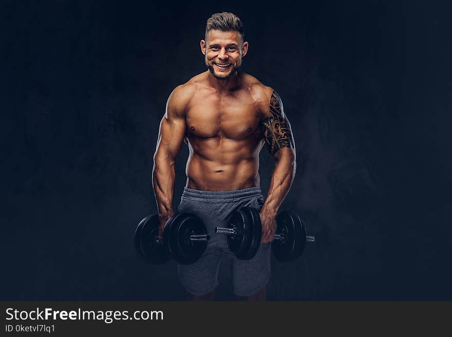 Smiling stylish bodybuilder with a tattoo on his arm, doing the exercises with dumbbells. Isolated on a dark background. Smiling stylish bodybuilder with a tattoo on his arm, doing the exercises with dumbbells. Isolated on a dark background.