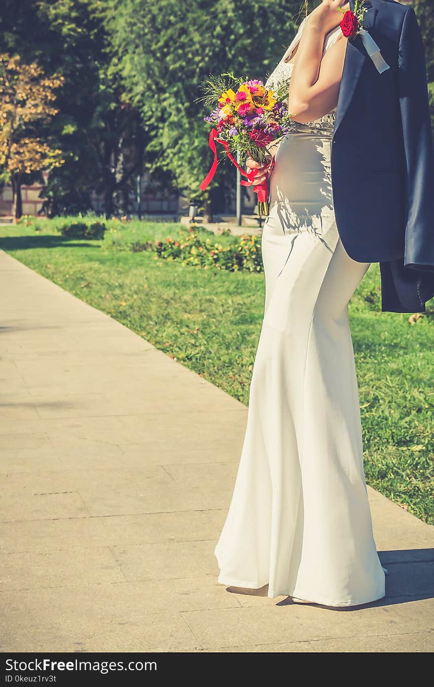 Elegant bride in a white long wedding dress holding groom`s jacket over her shoulder and colorful wedding bouquet of pink and yellow flowers. Vintage filter effect. Copy space. Elegant bride in a white long wedding dress holding groom`s jacket over her shoulder and colorful wedding bouquet of pink and yellow flowers. Vintage filter effect. Copy space.