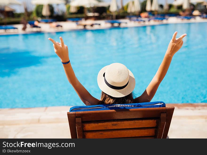 Rear view of Young beauty Woman in straw hat relaxing with hands up on chaise-lounge by the pool