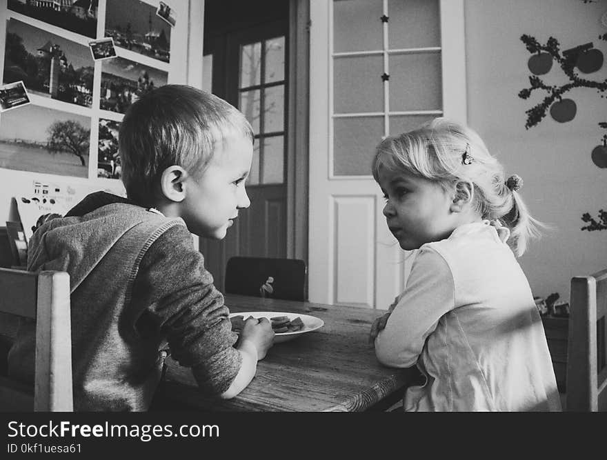 Grayscale Photo of Boy and girl sitting on a dining table chairs