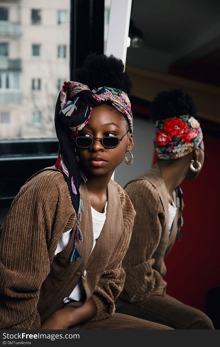 Woman Wearing White Shirt and Brown Coat With Sunglasses and Floral Turban Taking Selfie With Mirror Beside Her