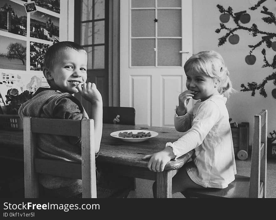 Grayscale Photo of Two Kids Sitting on Dining Table chairs